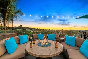 An outdoor seating area with a round wooden table, multiple sofas with blue cushions, and a fire pit in the center.