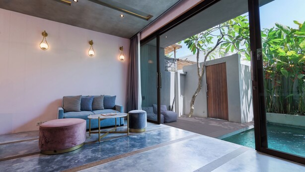 A modern living room with a sofa, ottoman, and coffee table, a glass door leading to a pool, and a view of greenery outside. - Liminal Palace Villa 1 by Betterplace (Canggu)