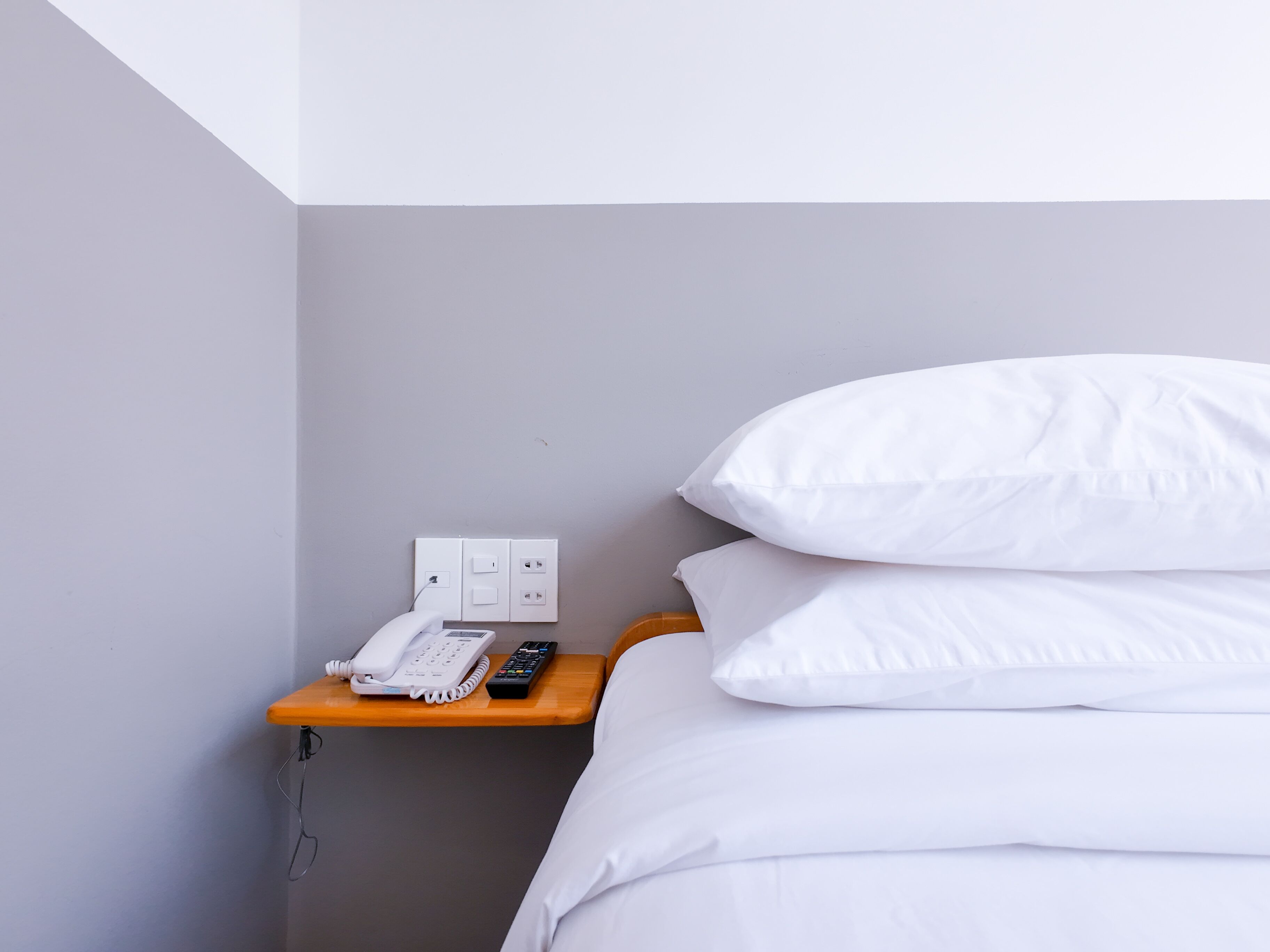 A hotel room with a bed, white pillows, a bedside table with a phone and remote control, and a wall-mounted light switch.