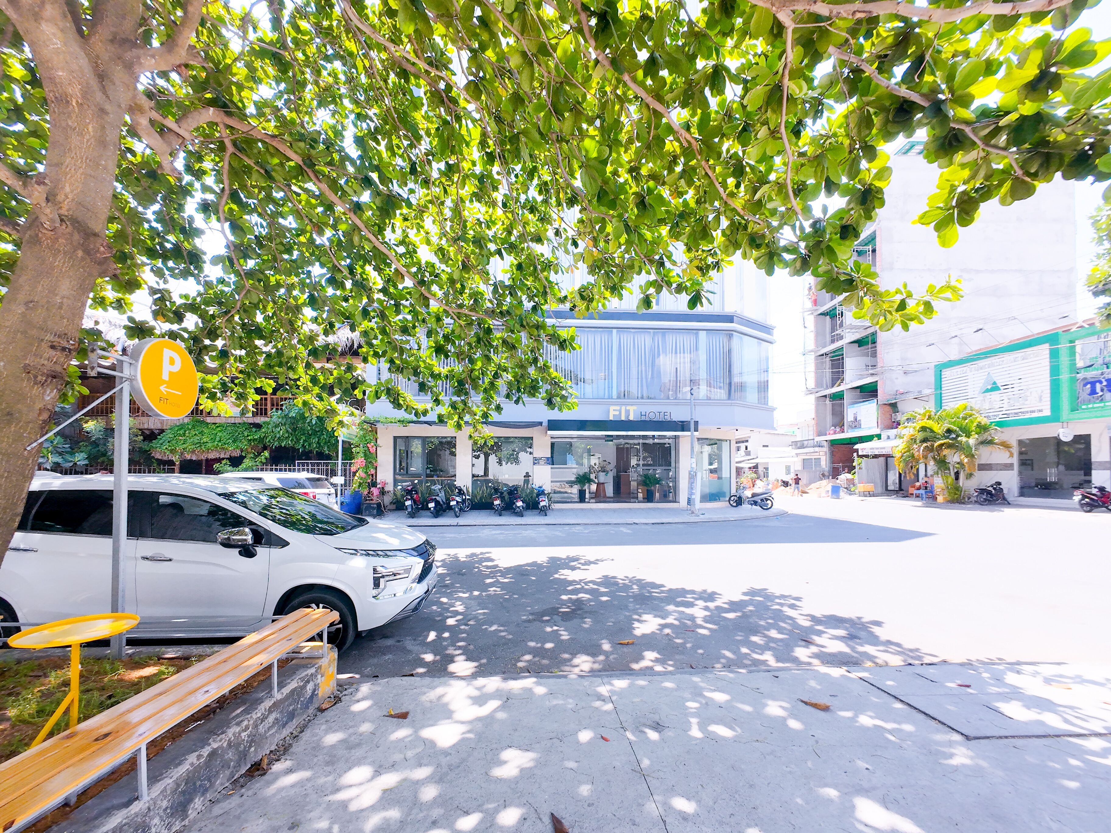 A hotel with a modern facade, a parking area, and a street with parked cars and a bench.