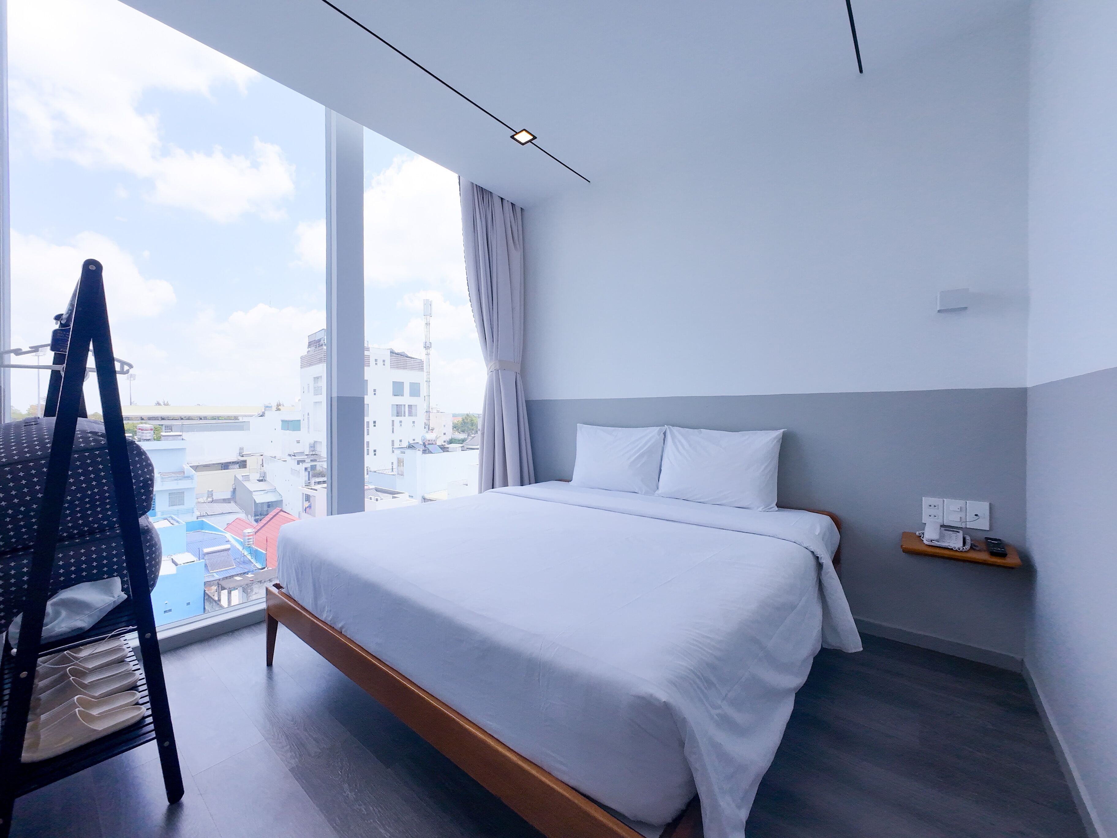 A hotel room with a large bed, a chair with a star-patterned bag, a small table with a phone, and a view of buildings through large windows.