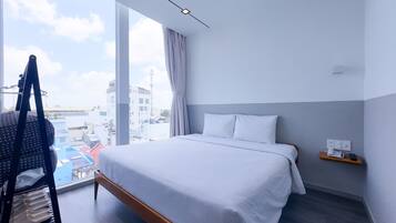 A hotel room with a large bed, a chair with a star-patterned bag, a small table with a phone, and a view of buildings through large windows.