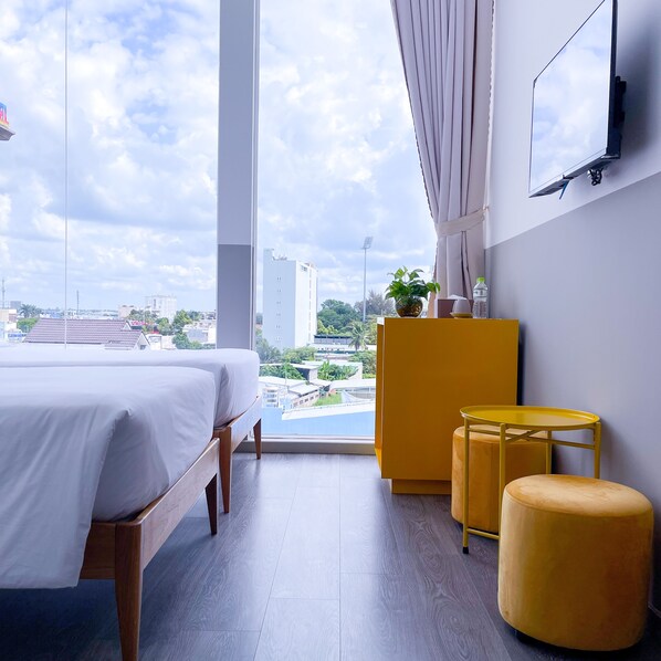 A modern hotel room with a large bed, a small table, a yellow ottoman, and a view of the cityscape through the window. - FIT Hotel Can Tho (Can Tho)