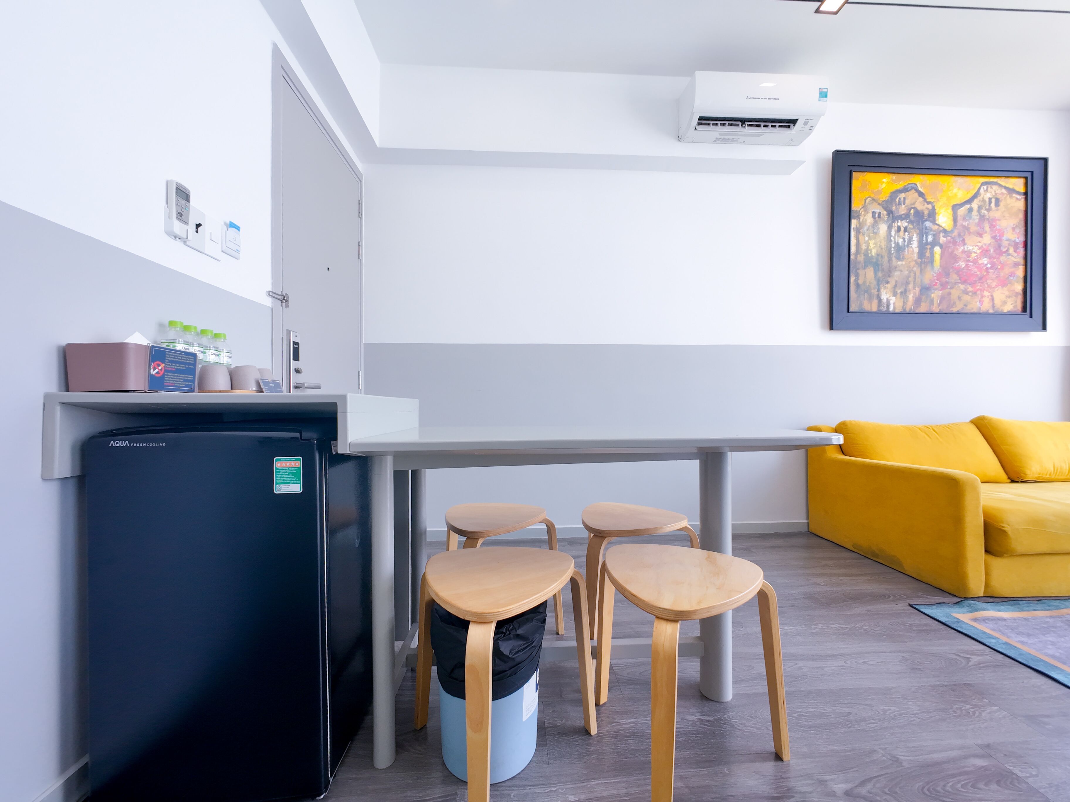 A modern kitchen with a refrigerator, a sink, and a dining area with stools. There is a yellow sofa and a painting on the wall.