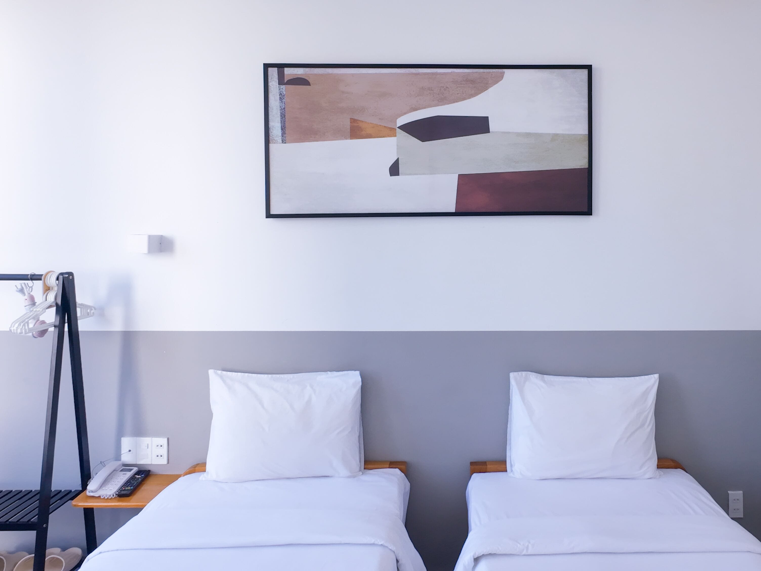 A hotel room with two beds, a painting on the wall, a phone on a bedside table, and a shoe rack.