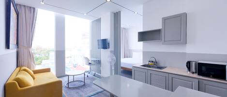 A modern hotel room with a sofa, dining table, kitchen area, and a large window offering a city view.