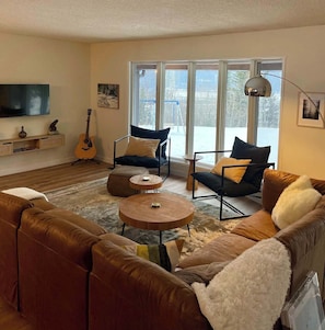A living room with a large window, a flat-screen TV, a guitar, and a sofa. - Nell’s Mountain Lodge - Movie Theatre|Arcade|Hottub|Fire Pit|18minKicking Horse (Golden)