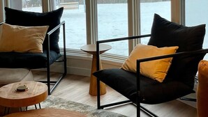 Una habitación con ventanas amplias, una silla de metal negro con cojines, una mesita de madera y una lámpara colgante.