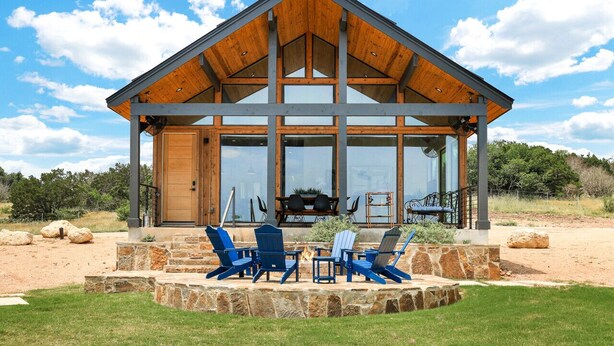 A modern lodge with a large glass facade, a stone patio with outdoor seating, and a wooden roof structure. - Cozy, Hill Country Cabin! (New Braunfels)