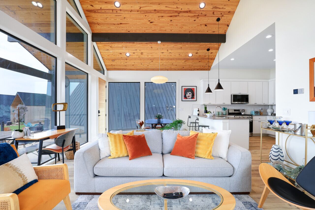 A modern living room with a grey sofa, orange and yellow cushions, a round coffee table, and a wooden ceiling.