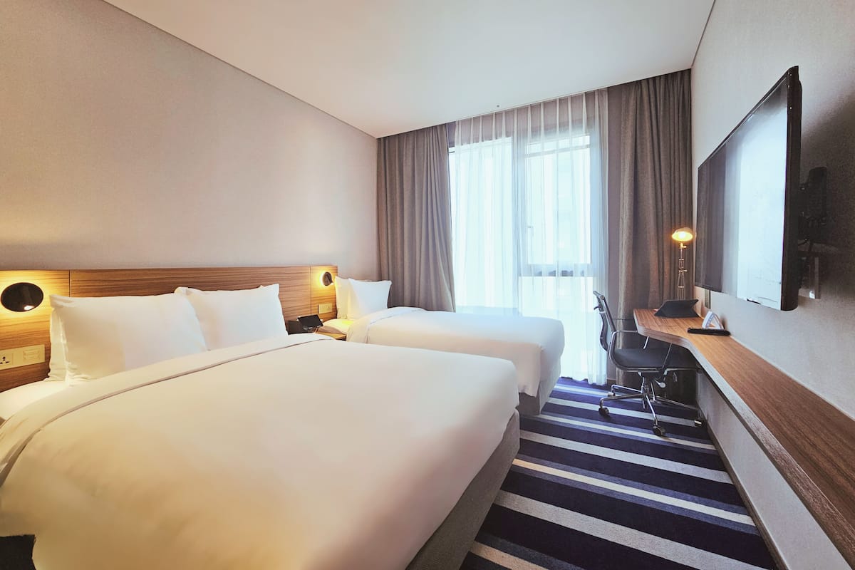 A hotel room with two beds, a desk with a chair, a large mirror, and a window with curtains.