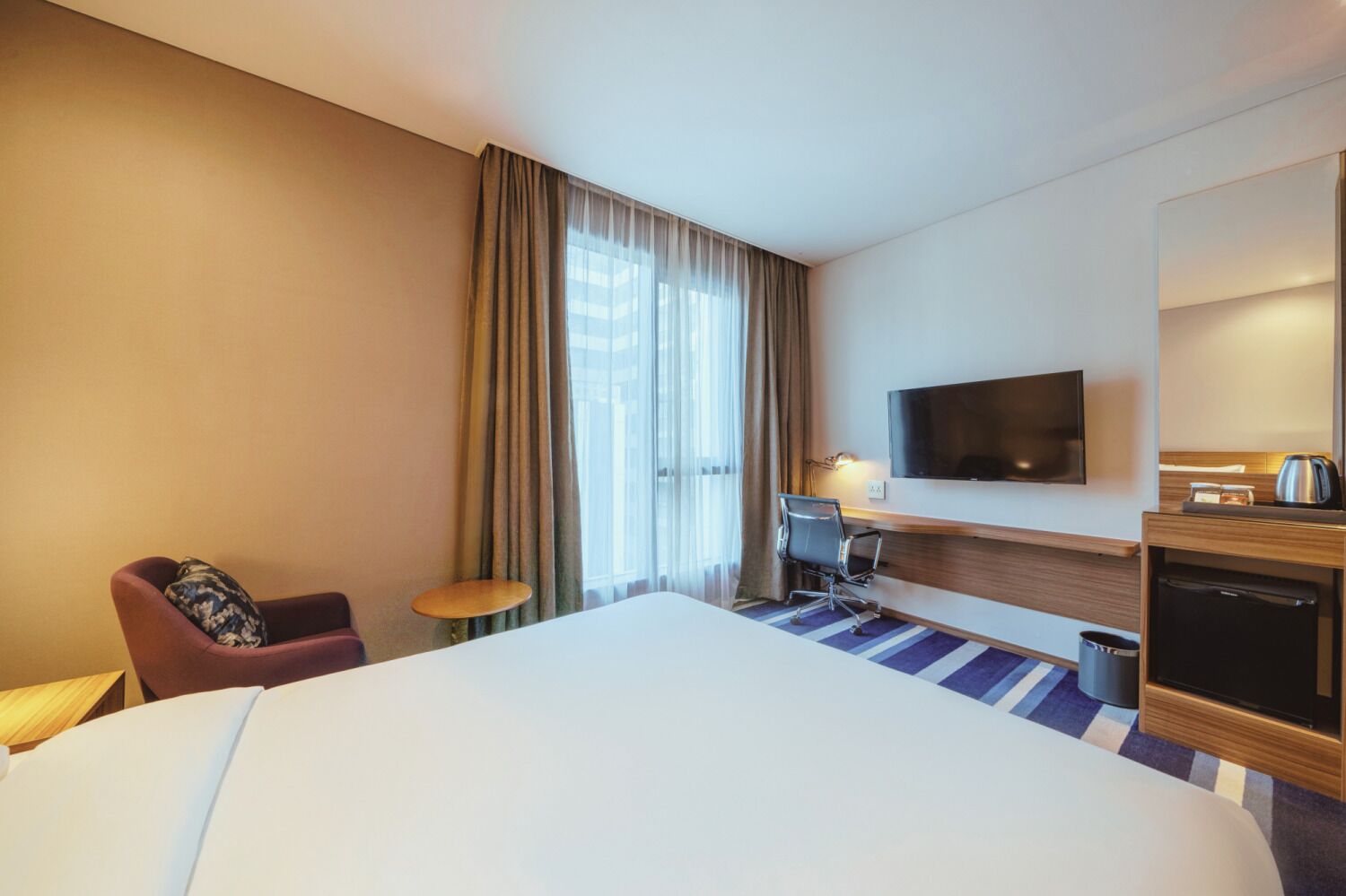 Habitación de hotel con una cama grande, un escritorio con silla, televisión y una kitchenette pequeña.