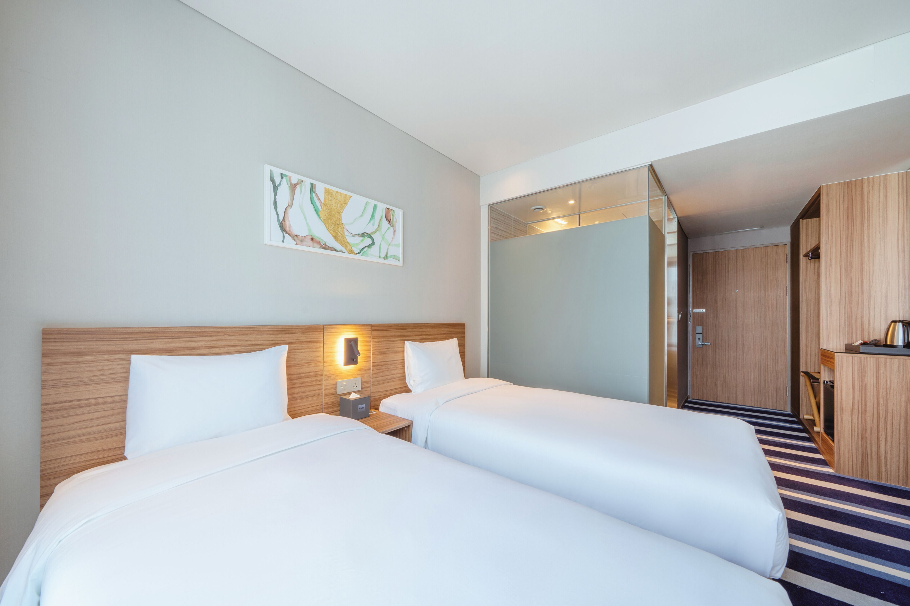 Une chambre d’hôtel avec deux lits, une tête de lit en bois, une table de chevet avec une lampe et un tableau au mur.