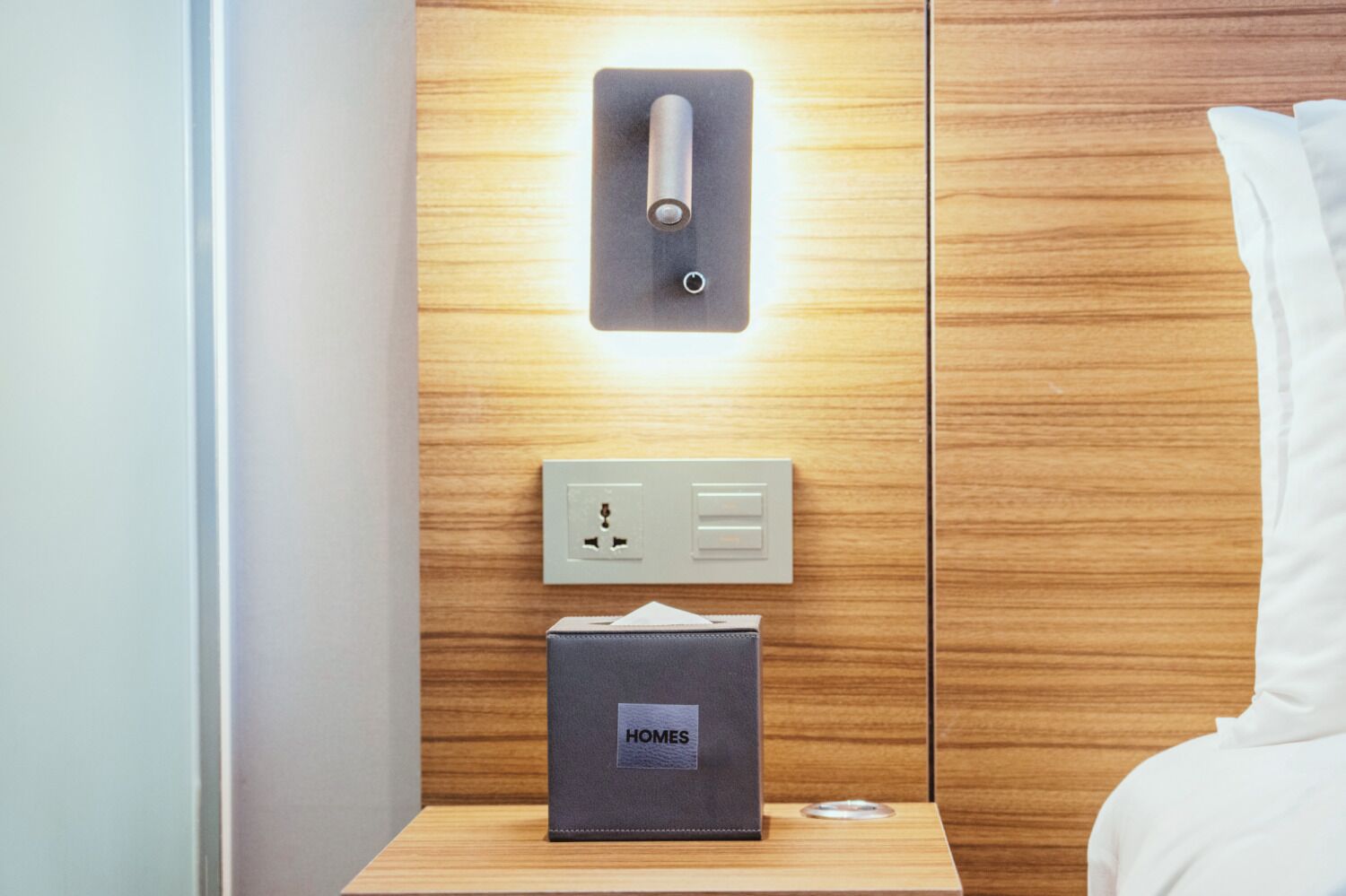 A hotel room with a wooden panel wall, a bedside table with a lamp, a power outlet, and a small box labeled "HOMES".