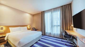 A hotel room with a large bed, a desk, a chair, a TV, and a window with curtains.