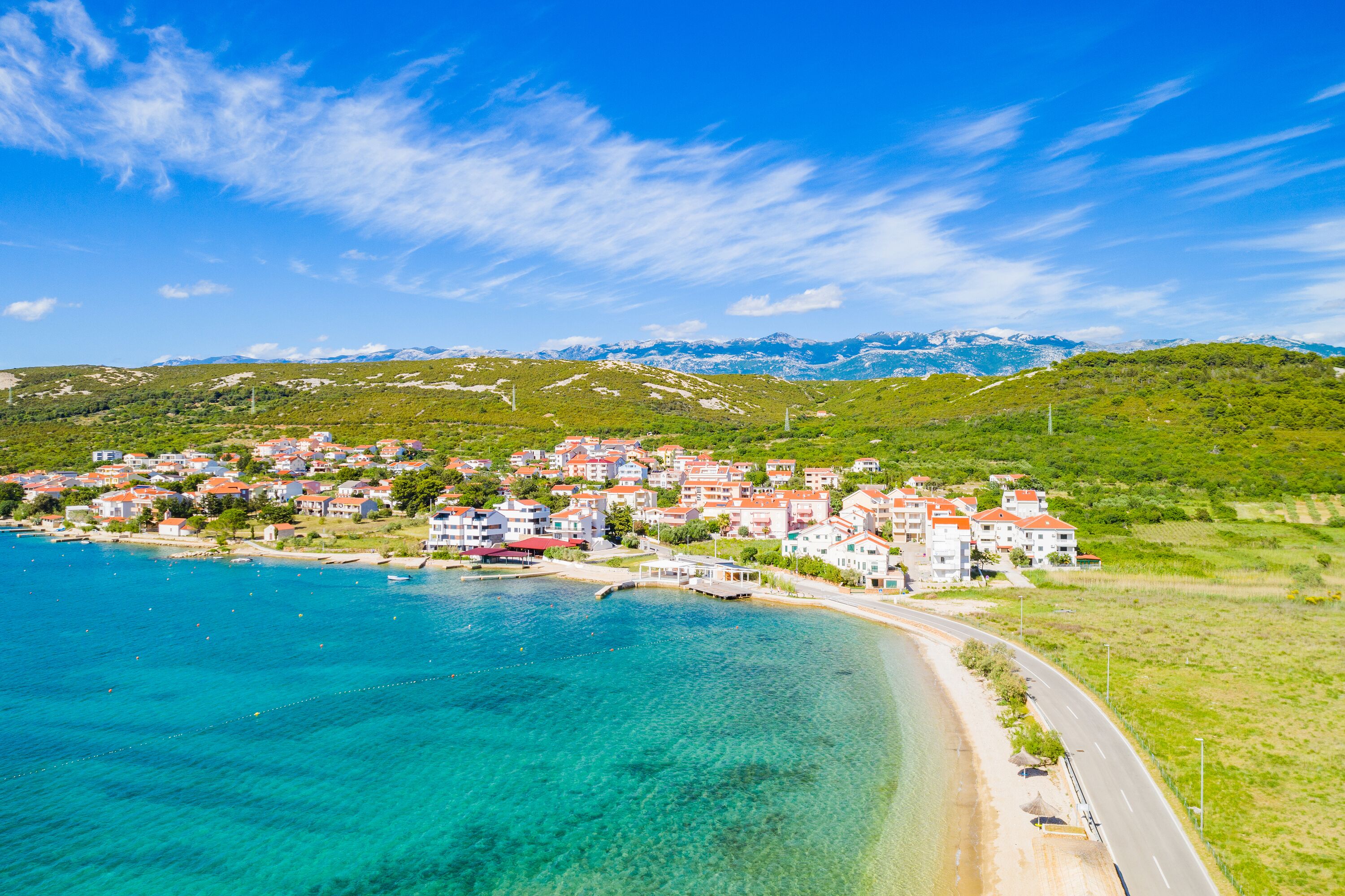 A coastal town with a clear blue sea, a road along the shore, and buildings with red roofs.