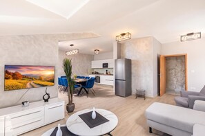 A modern living room with a flat-screen TV, a dining area with blue chairs, a kitchen with a stainless steel refrigerator, and a grey sofa. - Family Resort Santa Maria II Penthouse (Novalja)