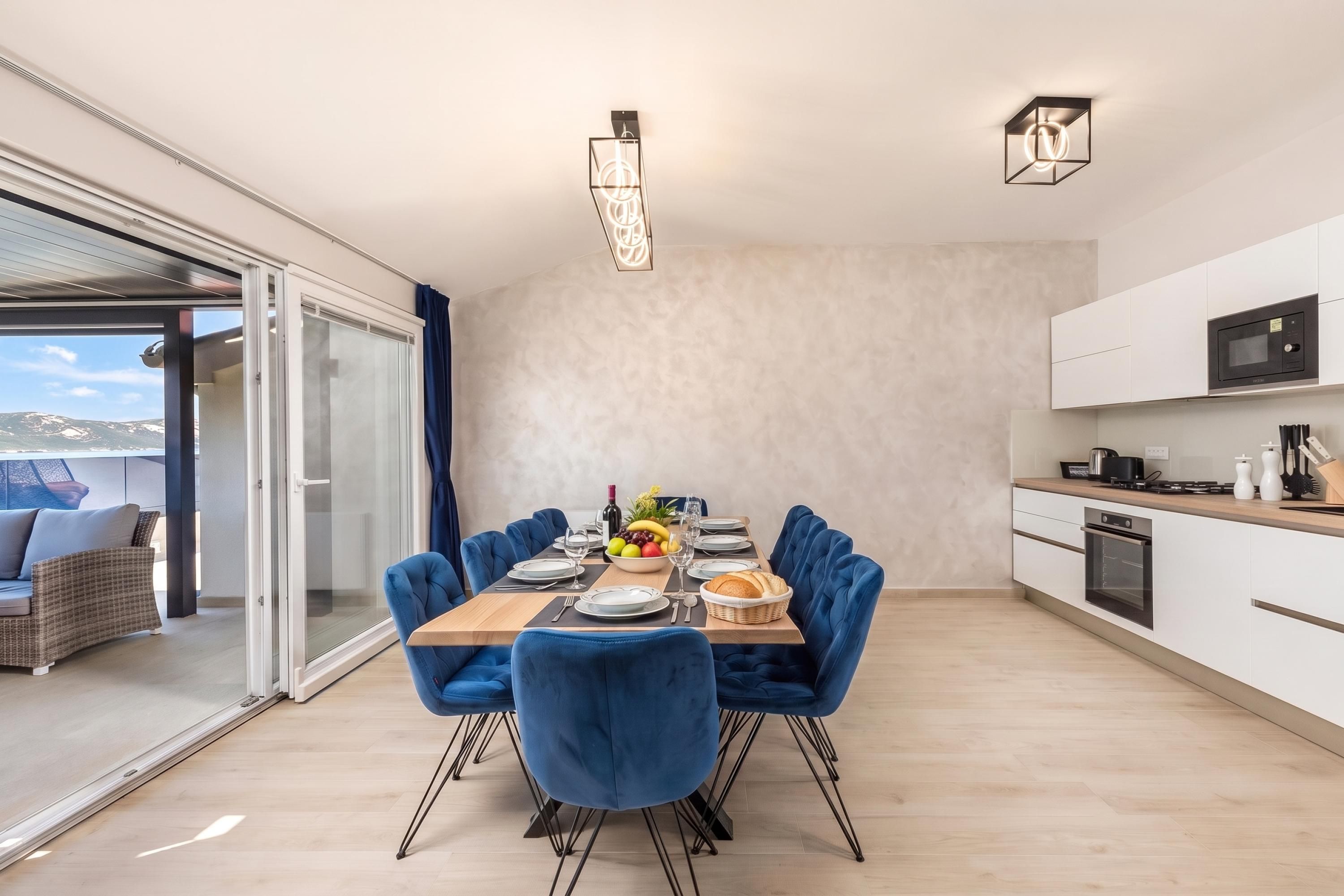 Ein moderner Essbereich mit einem Holztisch, blau gepolsterten Stühlen und Blick durch eine Glastür nach draußen.