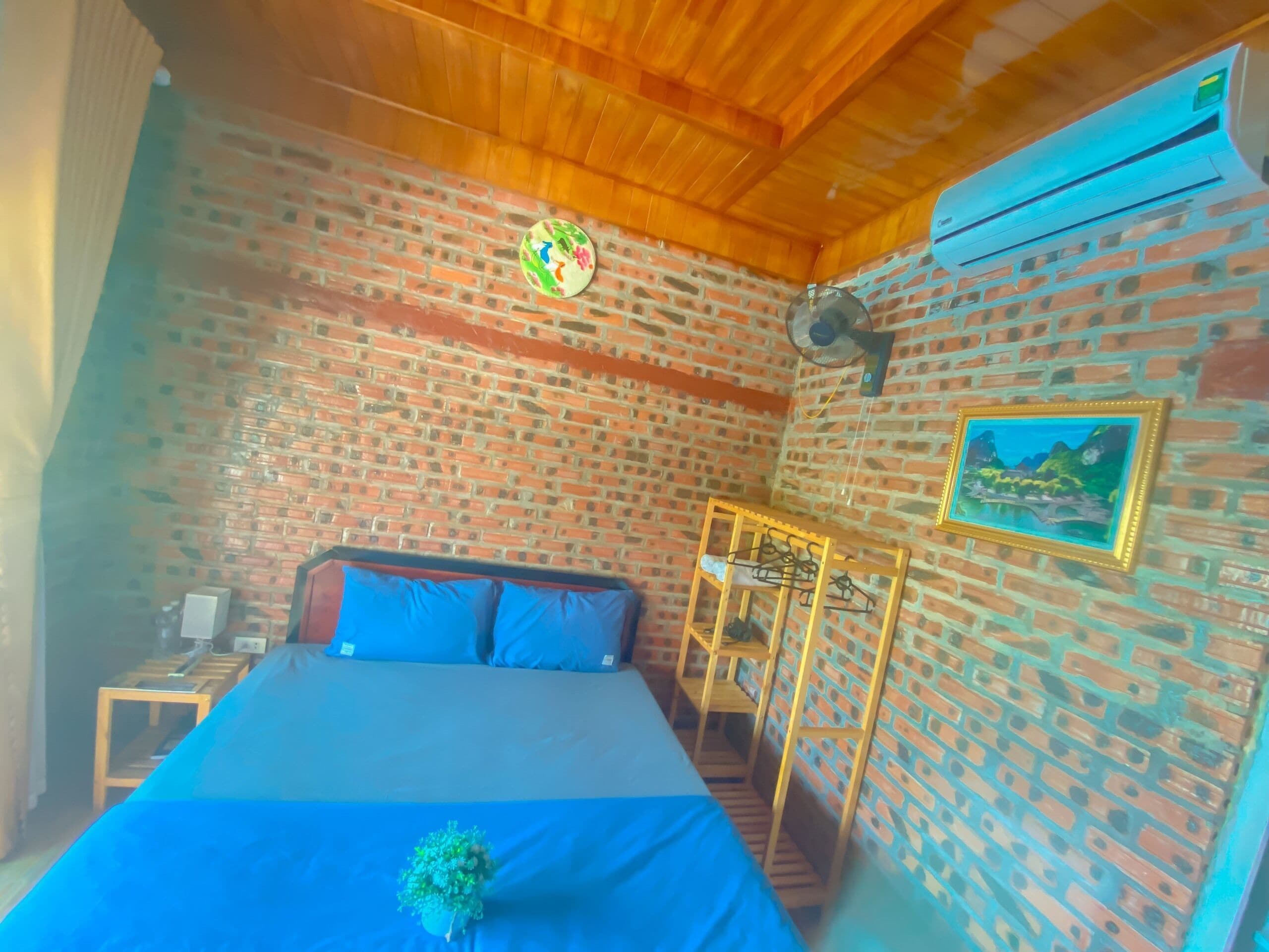 A room with a bed, a wooden headboard, a brick wall, a ceiling fan, a small table, and a picture frame.