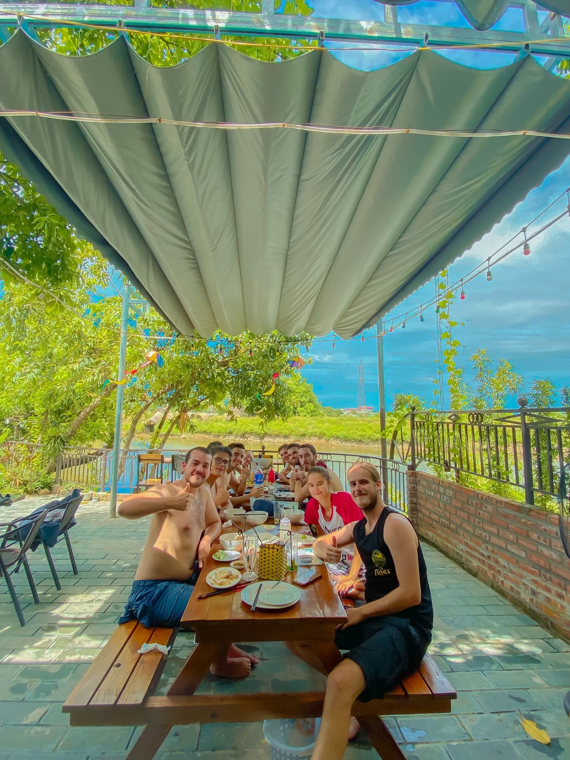 A group of people dining at an outdoor table under a canopy, with a view of greenery and a body of water in the background.