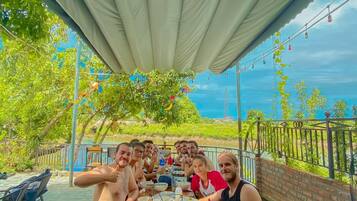 A group of people dining at an outdoor table under a canopy, with a view of greenery and a body of water in the background.