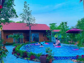 A swimming pool area with a red-roofed building, surrounded by tropical plants and a tiled edge. - D&D Riverside Homestay (Hoa Lu)