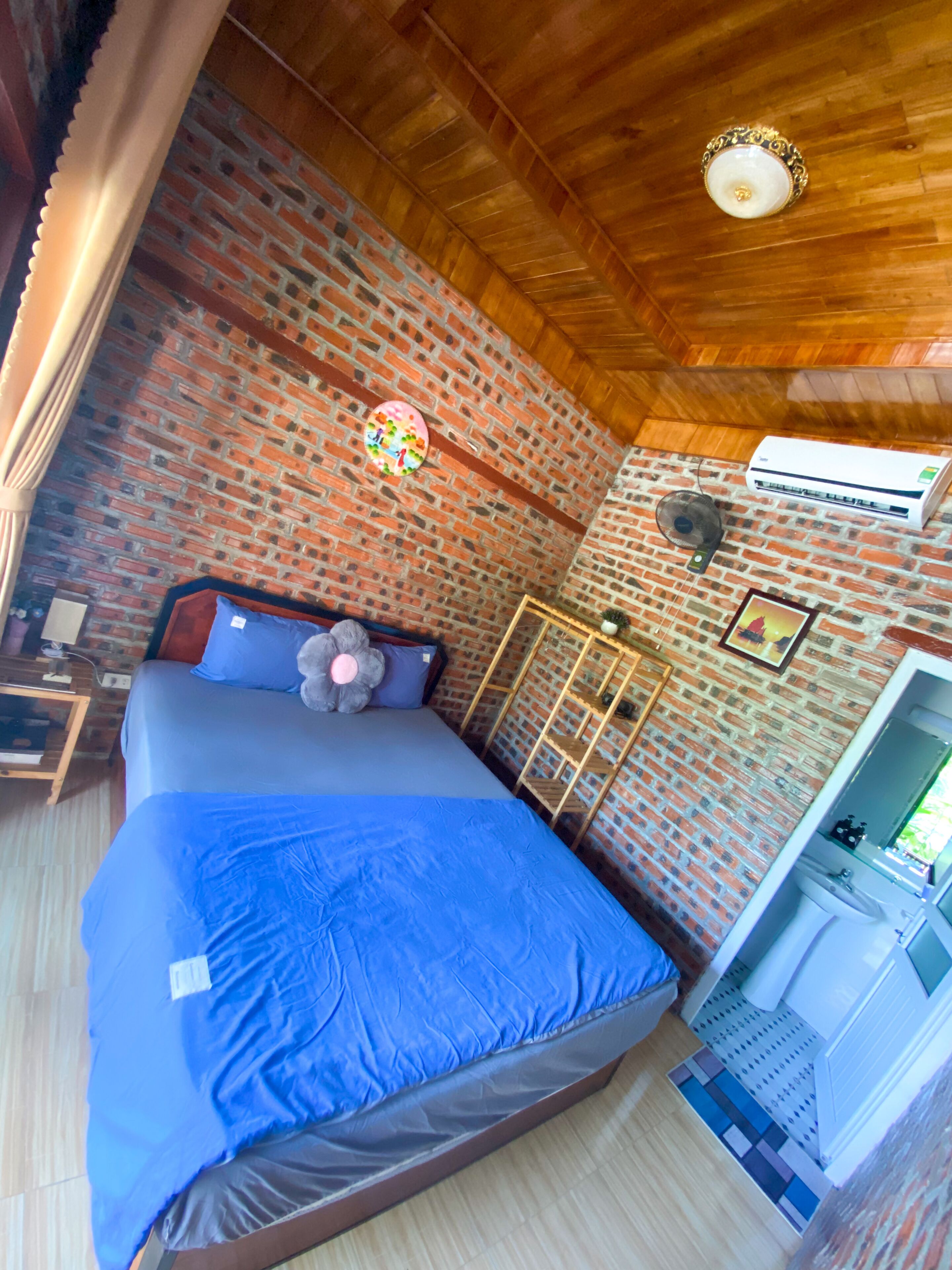 Una habitación con pared de ladrillo, una cama con colcha azul, un ventilador y un equipo de aire acondicionado.