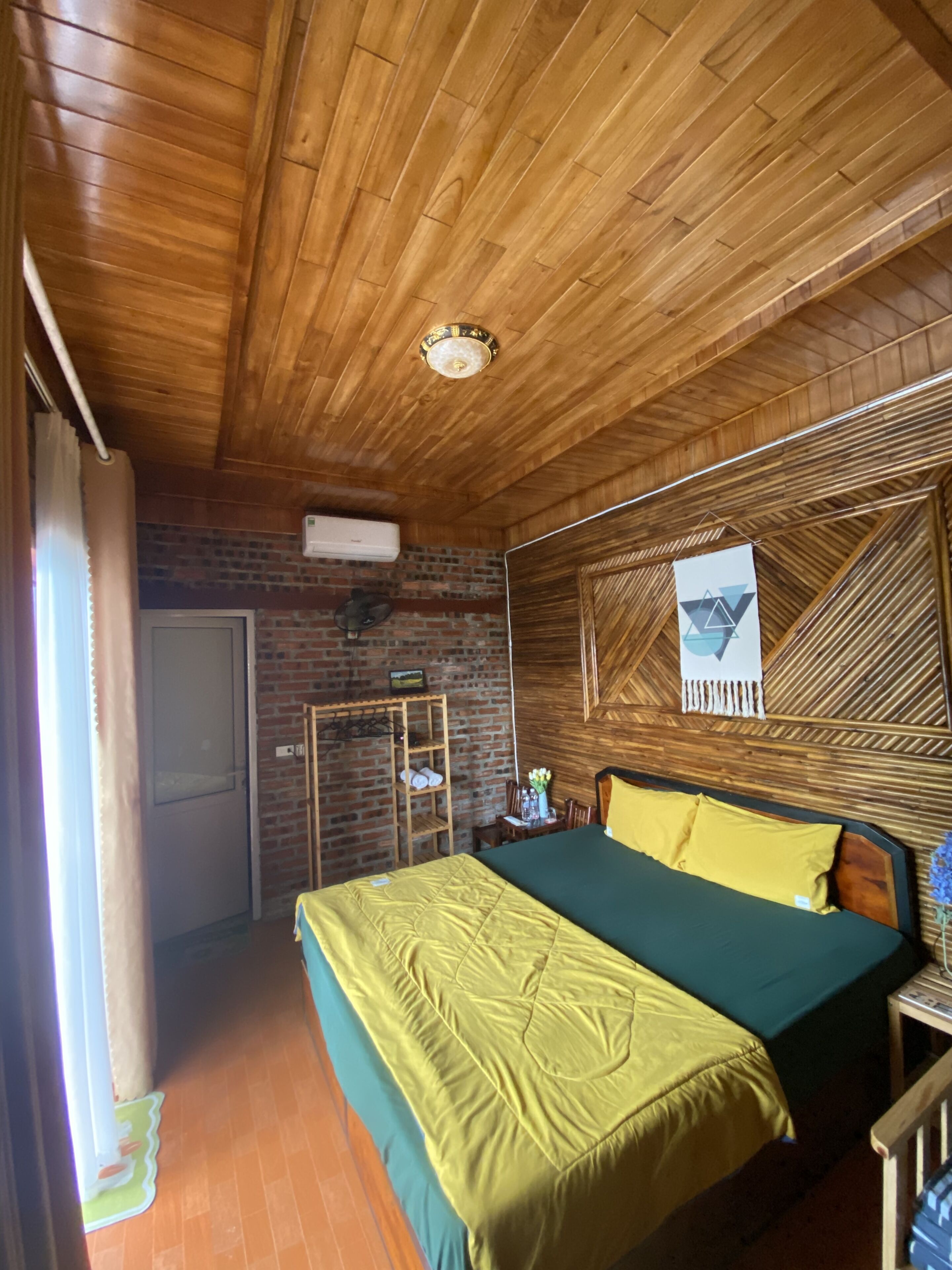 A room with a wooden ceiling, a bed with green and yellow bedding, a brick wall, and a window with curtains.