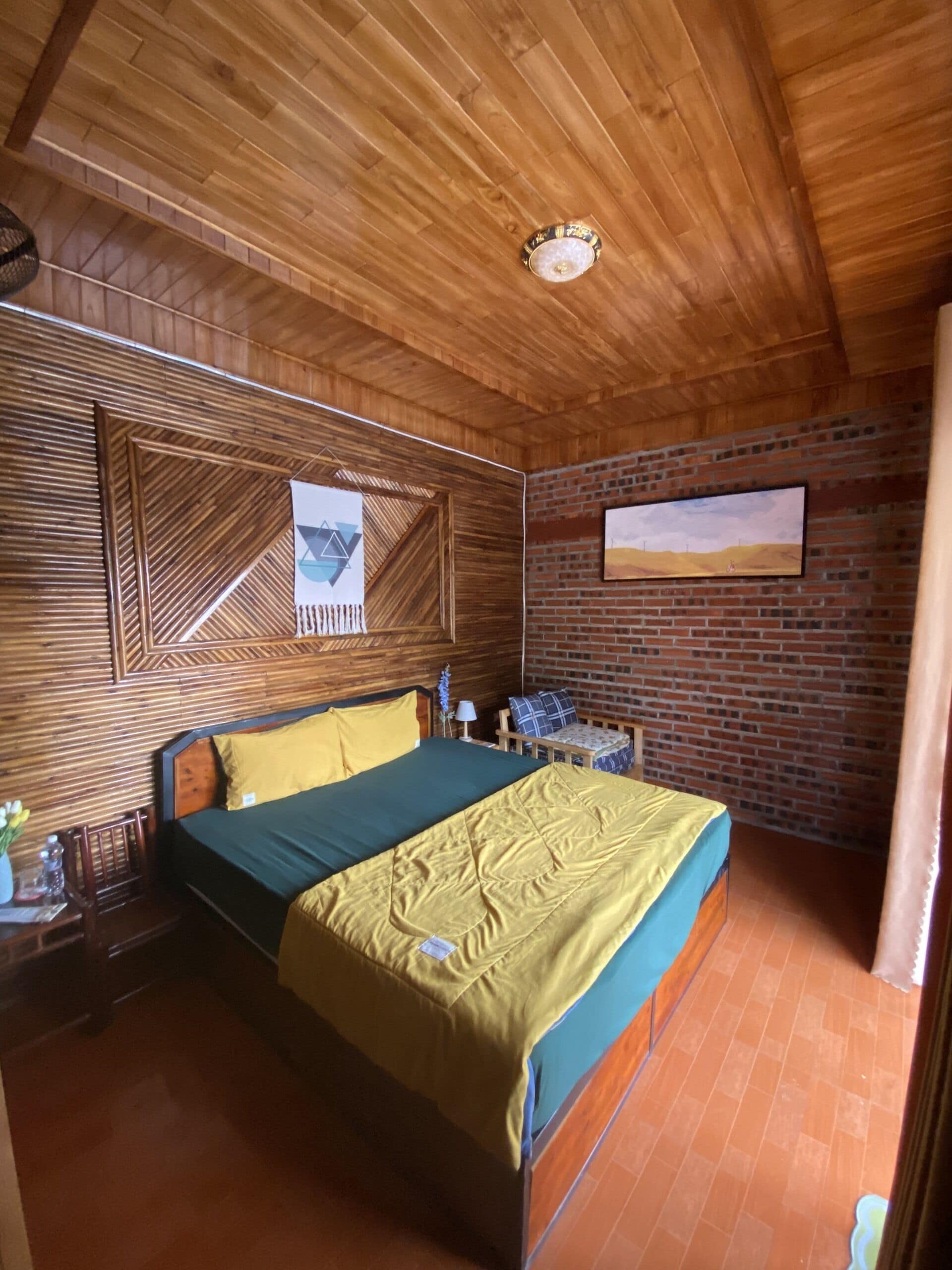 Una habitación con techo de madera, cama con ropa de cama verde y amarilla, pared de ladrillo y un cuadro enmarcado.