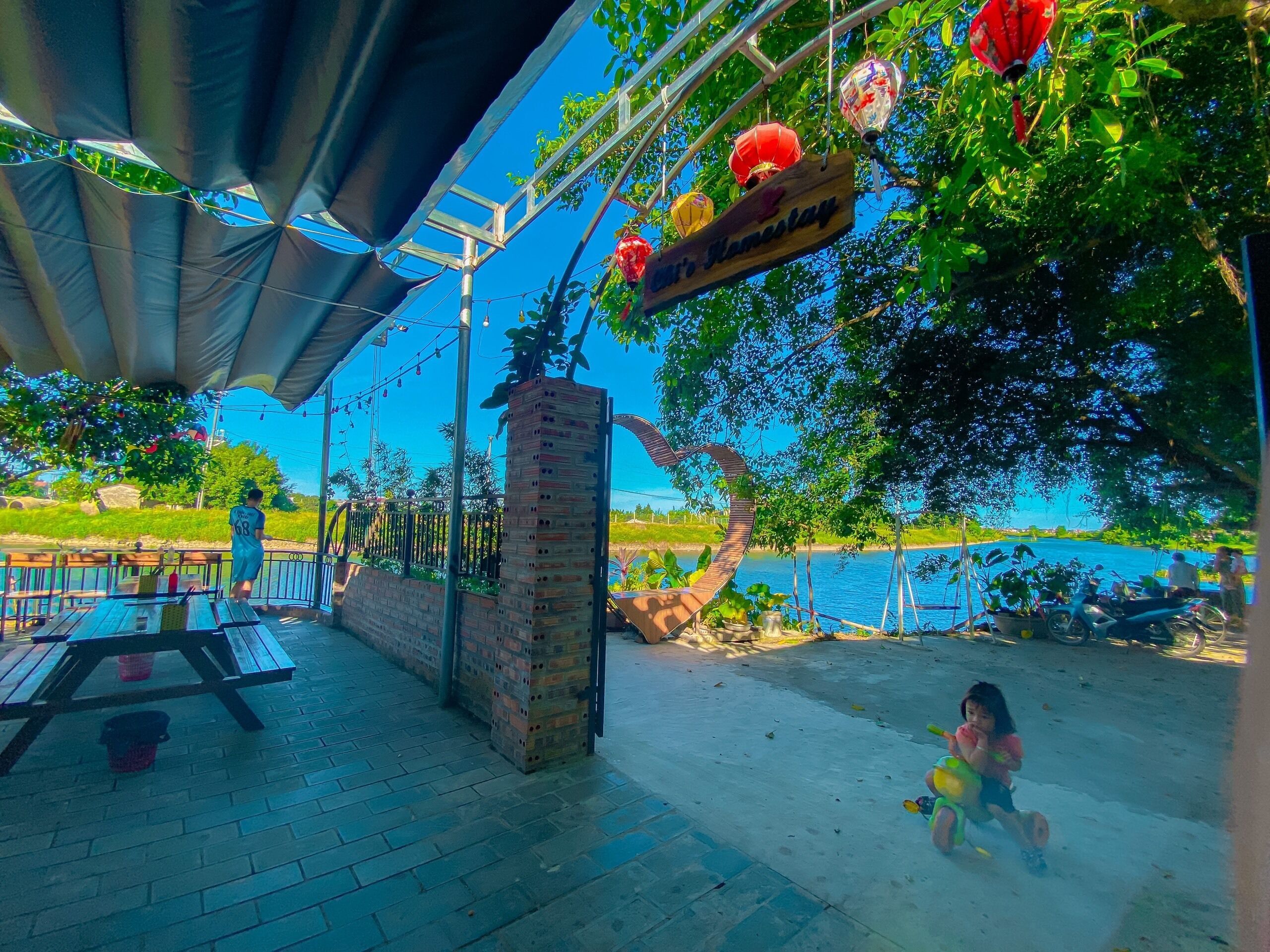 An outdoor seating area with a brick structure, a sign reading "M's Flamboyance," and a view of a body of water.