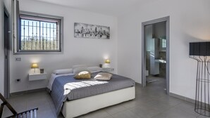 A bedroom with a bed, bedside lamps, a window with bars, a cityscape painting, and a bathroom visible through an open door. - Villa Delle Ginestre 9 2 Emma Villas (Cingoli)