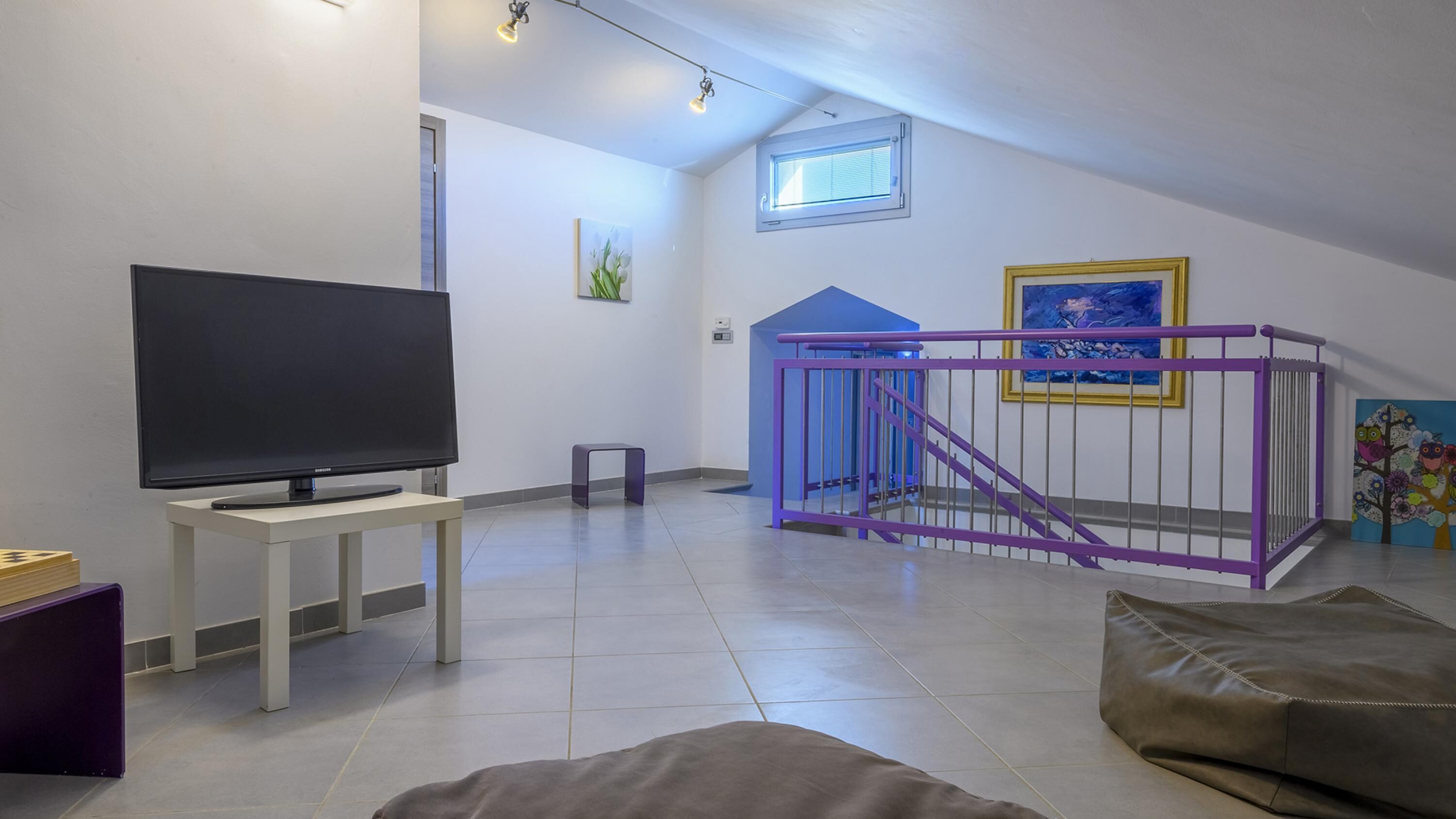 A living room with a flat-screen TV on a white stand, a purple playpen, and a blue staircase.