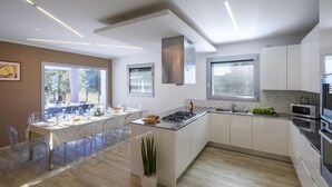 A modern kitchen with white cabinets, a central island, and a dining area with clear chairs. - Villa Delle Ginestre 9 2 Emma Villas (Cingoli)