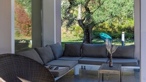 A modern living room with a sectional sofa, a small table, and a view of a garden with a tree and a chair. - Villa Delle Ginestre 9 2 Emma Villas (Cingoli)