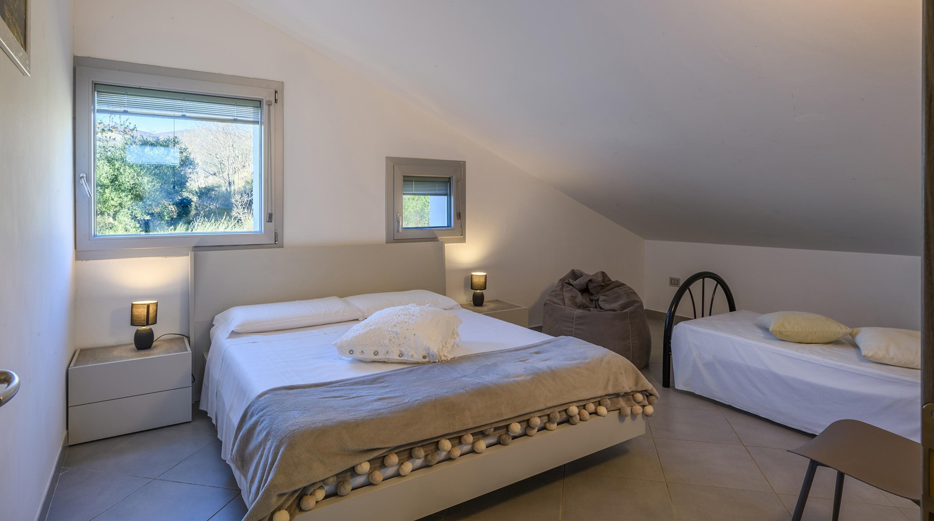 A bedroom with two beds, a window, and a small bedside table with a lamp.