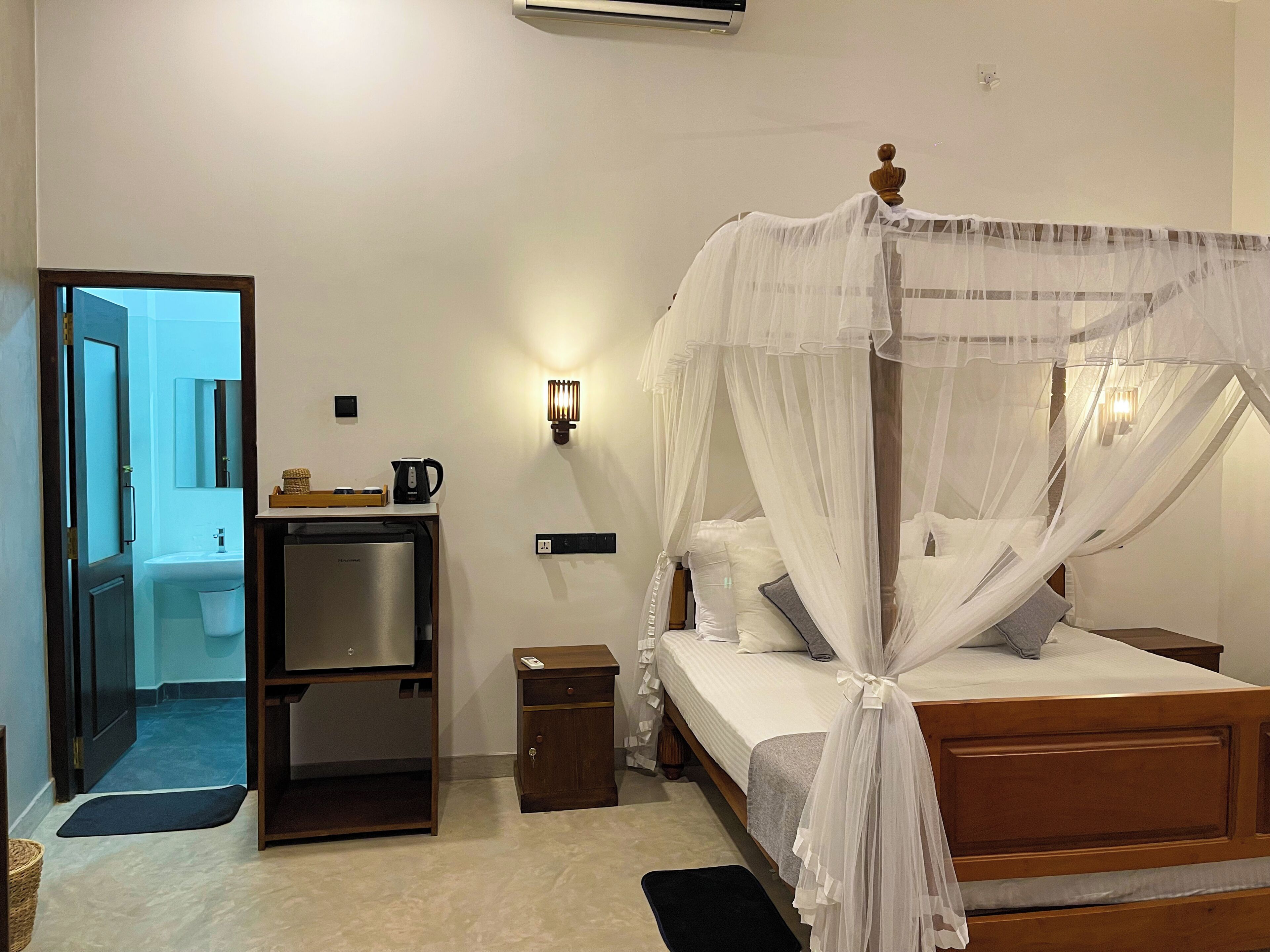 A bedroom with a canopy bed, a bedside table, a nightstand, a desk with a chair, a mirror, and a wall-mounted light fixture.