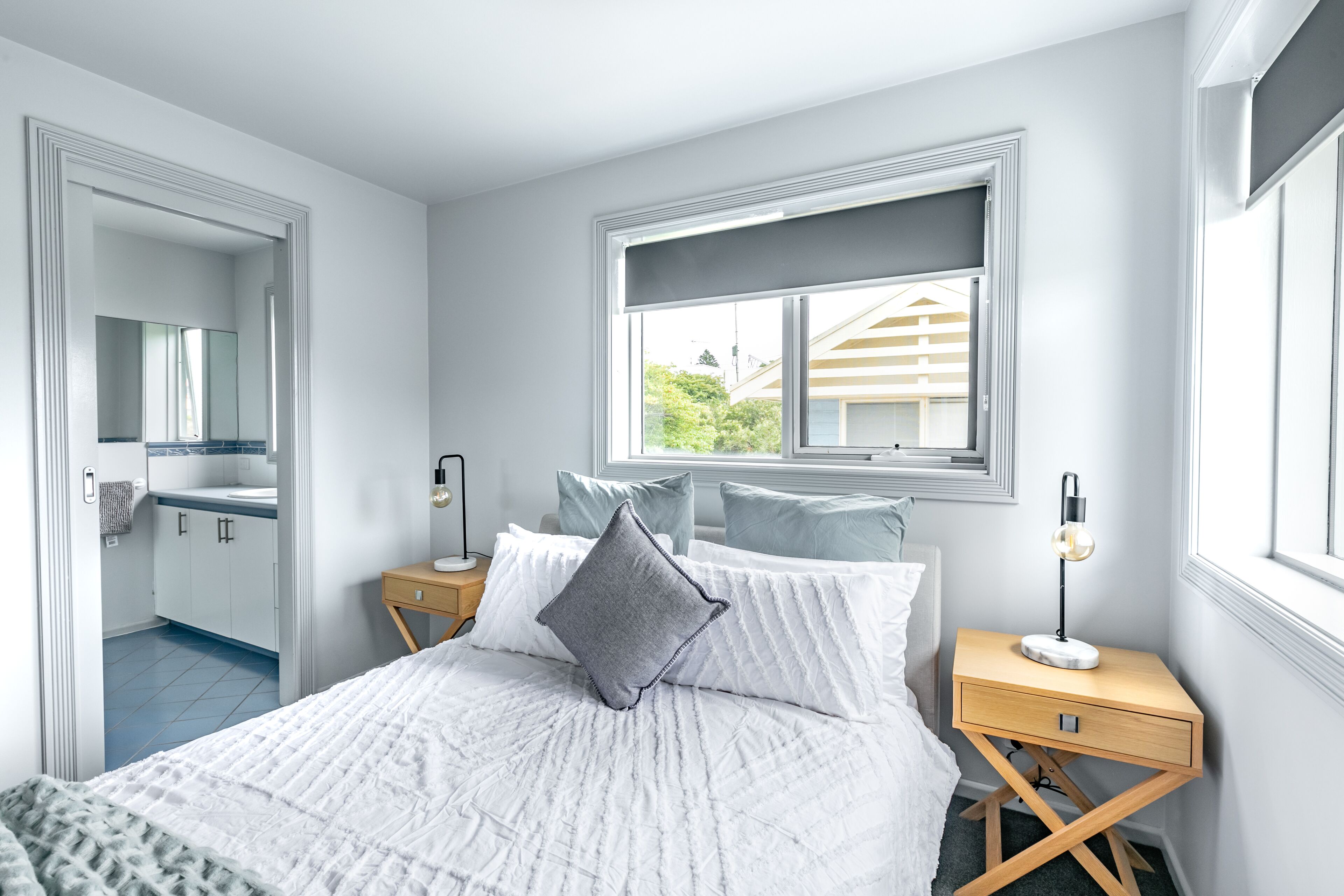 Une chambre à coucher avec un lit, une table de chevet, une fenêtre et une salle de bain visible à travers une porte ouverte.