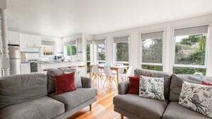 A spacious living area with grey sofas, a dining table, and large windows offering a view of the outdoors. - Ocean Breeze: 100m from the beach. Pet Friendly (Ocean Grove)