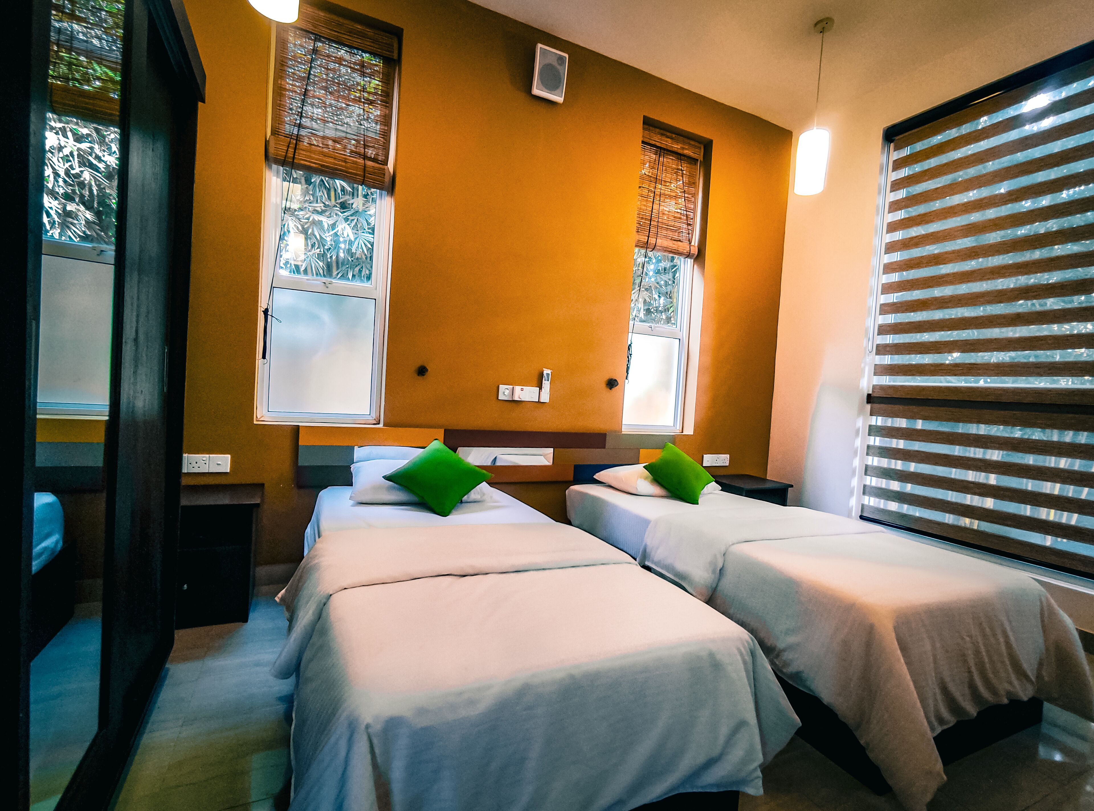 A hotel room with two beds, each with a green pillow, a window with blinds, and a wall-mounted air conditioning unit.