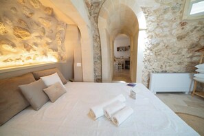 Ein Schlafzimmer mit Steinwand, einem Bett mit zusammengerollten Handtüchern und Blick zur Küche.
