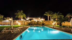 Ein Schwimmbad bei Nacht mit Liegestühlen und Palmen, umgeben von einer Steinmauer und einem überdachten Bereich mit Beleuchtung.