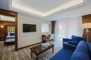 Quarto de hotel com cama, escrivaninha com computador, televisão, sofá e uma grande janela com cortinas.