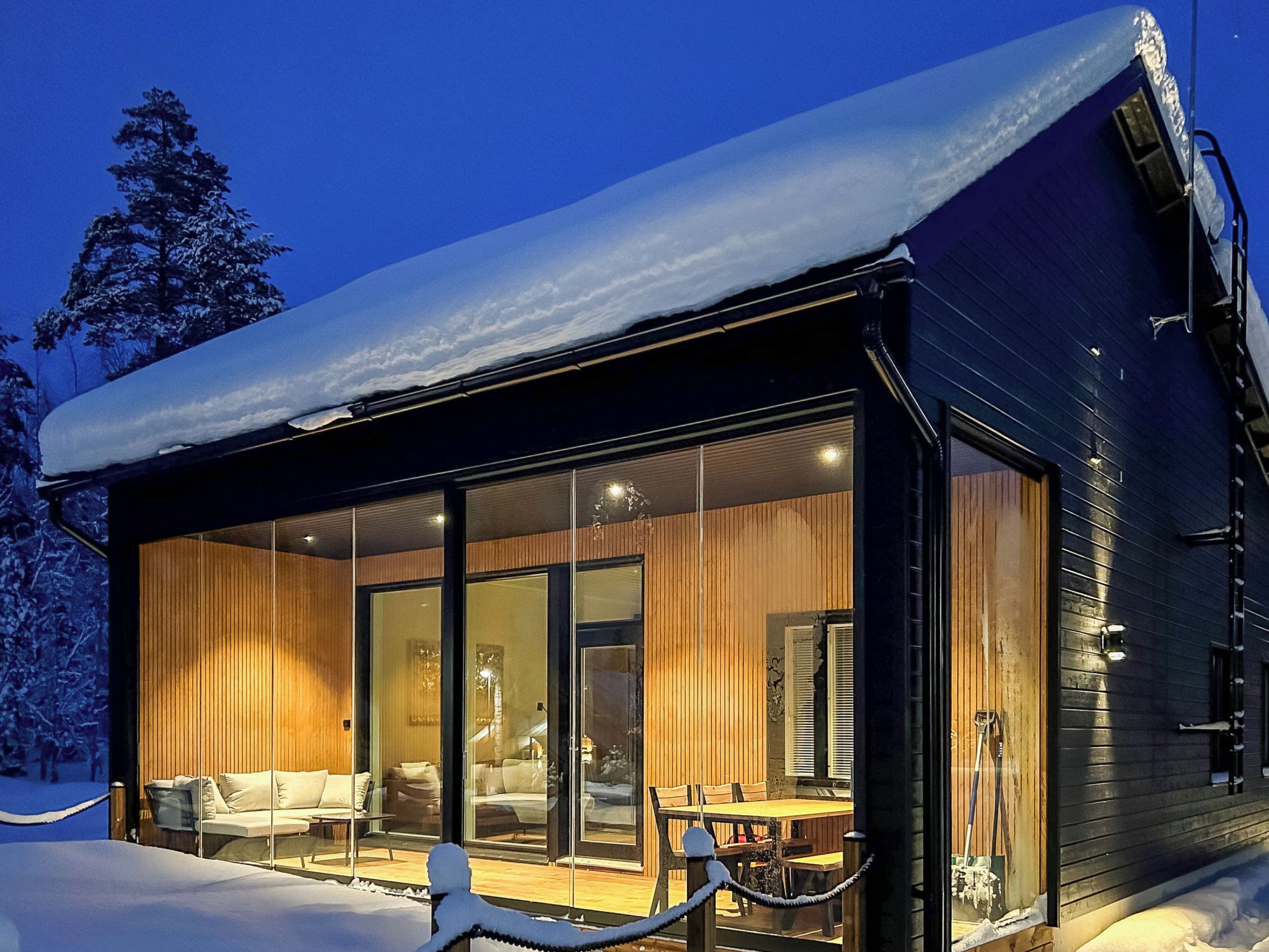 Una cabaña moderna con un espacio de estar con paredes de vidrio, interior de madera y un paisaje nevado.
