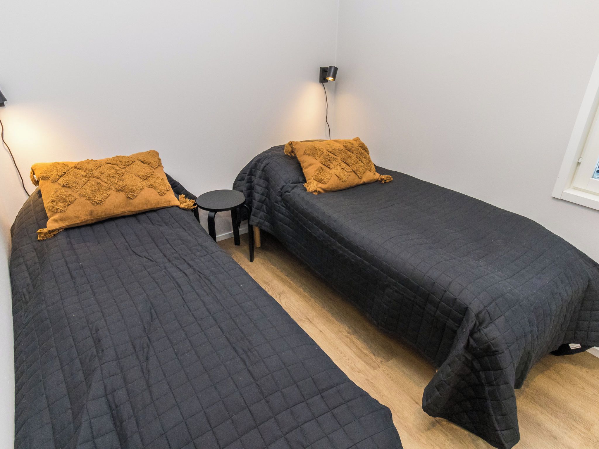 Une chambre avec deux lits simples, chacun recouvert d’une courtepointe matelassée de couleur sombre et agrémenté d’un coussin texturé de couleur moutarde. Une petite table ronde se trouve entre les lits.