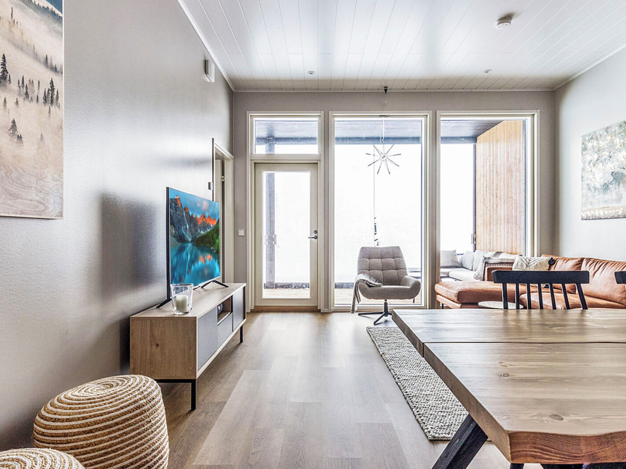 Un salon moderne avec une table à manger en bois, un canapé, un téléviseur posé sur un meuble et une grande fenêtre donnant sur l’extérieur.