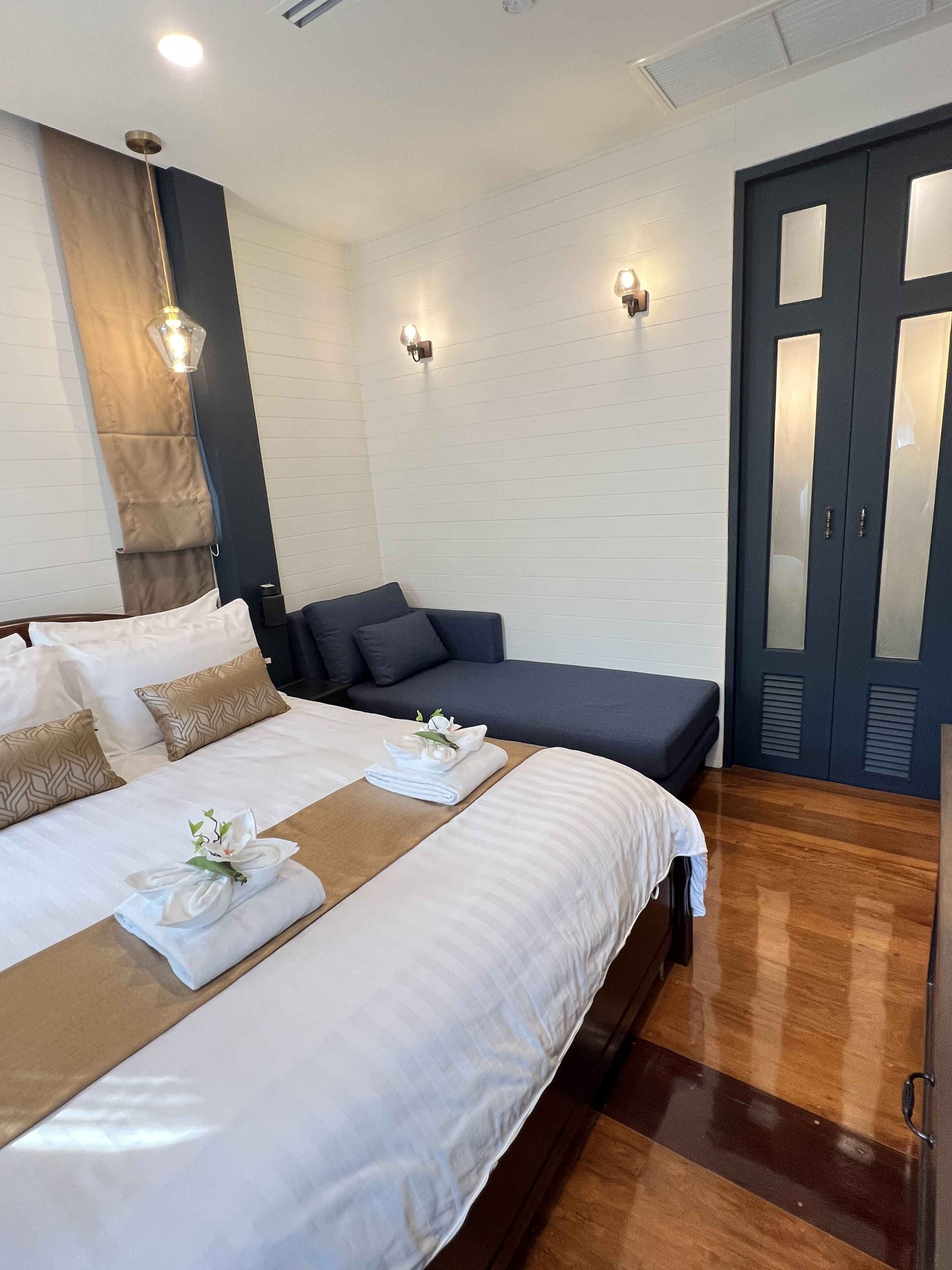 A hotel room with a large bed, a sofa, two wall-mounted lights, and a dark-colored wardrobe.