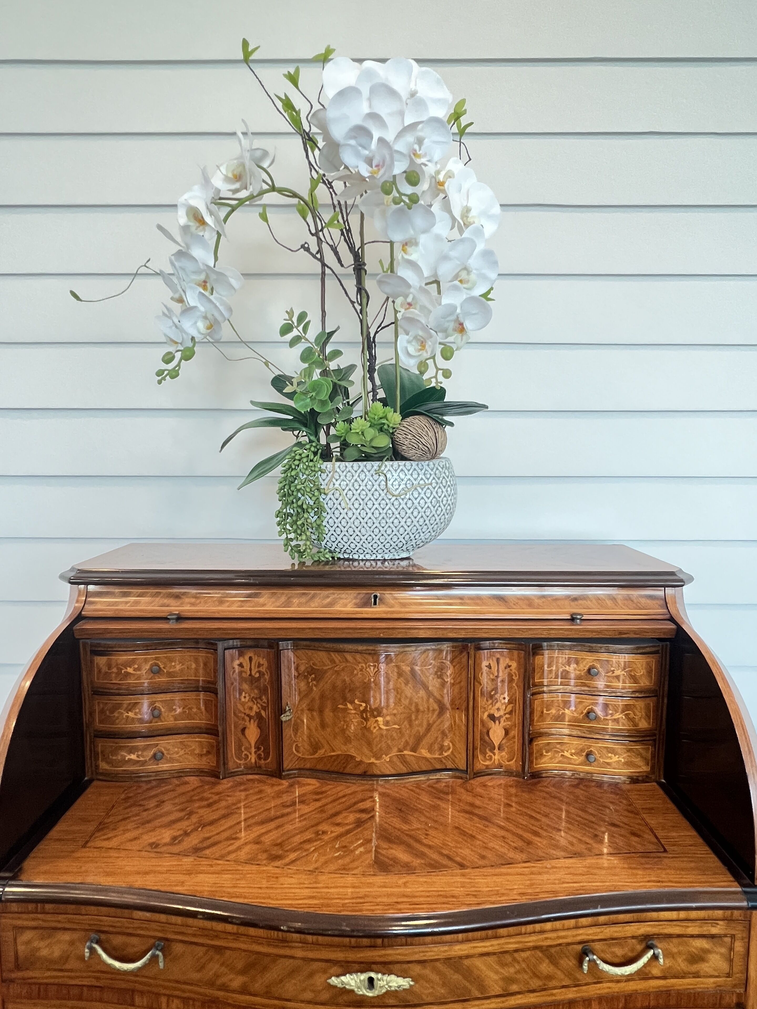 A wooden desk with a patterned vase containing white orchids and green foliage.