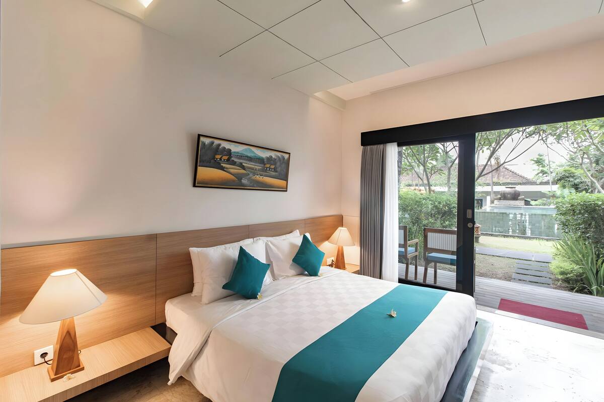 A hotel room with a large bed, a nightstand with a lamp, a painting on the wall, and a view of the outdoors.