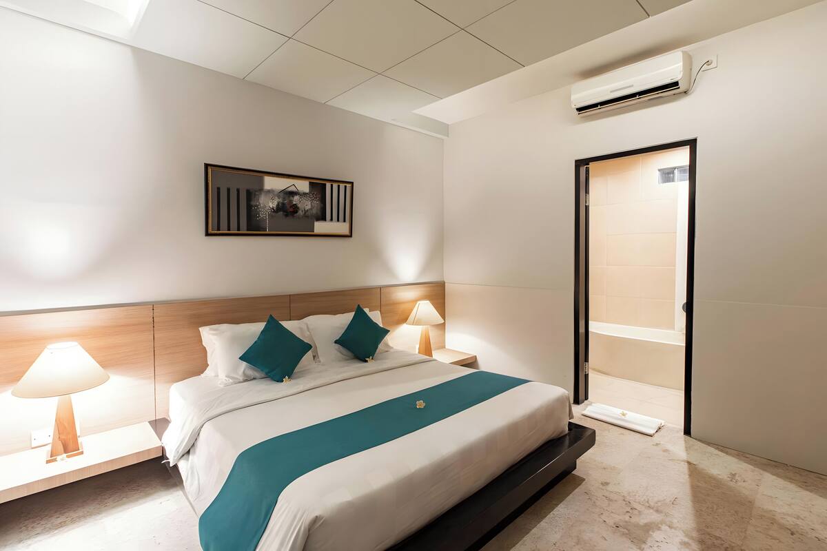 A hotel room with a bed, bedside lamps, a framed picture on the wall, an air conditioning unit, and a bathroom door.