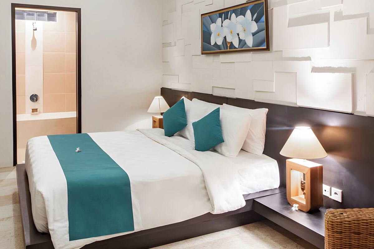 A hotel room with a bed, nightstands, a lamp, a painting, and a bathroom door.